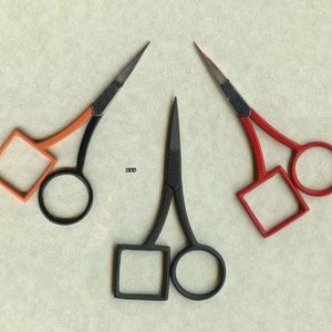 May include: Three pairs of small scissors with different colored handles. The scissors on the left have orange handles, the scissors in the middle have black handles, and the scissors on the right have red handles.