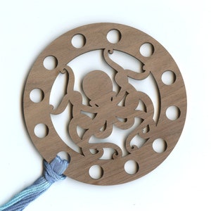 May include: A wooden octopus-shaped thread holder with 10 holes around the edge. A blue and light blue ombre thread is threaded through one of the holes.