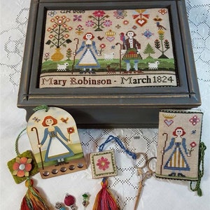 Può includere: Una scatola di legno con un disegno a punto croce che raffigura una coppia in un ambiente da giardino. La scatola è chiusa e ha un'etichetta che recita "Mary Robinson - Marzo 1824". La scatola è circondata da vari accessori per il cucito, tra cui un salvaguardia per aghi, un cuscinetto per aghi, un paio di forbici e un piccolo pezzo ricamato.