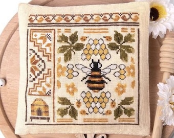 May include: Counted cross stitch pattern of a bee with honeycomb, flowers, and a beehive. The pattern is called "The Bee Quaker II" and is framed on a wooden surface.