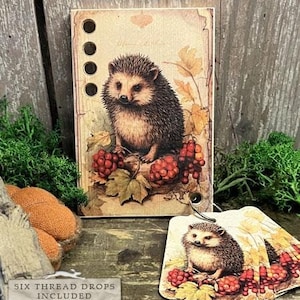 Könnte beinhalten: Ein Holz-Bastelset mit einem Igel, umgeben von roten Beeren und Herbstblättern. Das Set enthält sechs Garnfäden und ist Teil der Smart Thread Keeps-Kollektion. Der Text "HEDGEHOG" und "2024 NEEDLEWORK MARKETPLACE COLLECTION BY THE PRIMITIVE HARE" ist unten auf dem Bild gedruckt.