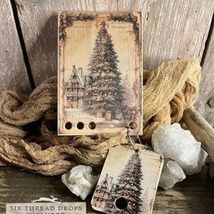 Può includere: Un ornamento per albero di Natale in legno in stile vintage con un'immagine dipinta di un albero di Natale e una casa. L'ornamento include sei gocce di filo e il testo "Smart Thread Keeps collection OH CHRISTMAS TREE 2024 NEEDLEWORK MARKETPLACE COLLECTION BY THE PRIMITIVE HARE" è stampato in basso.
