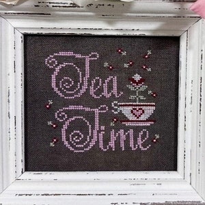 May include: A white framed cross-stitch with a pink and gray design that says "Tea Time" with a small teacup and flower design.