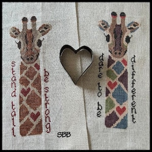 May include: Two cross-stitch patterns featuring giraffes with different colored patterns and text. The left giraffe says "Stand Tall Be Strong" and the right giraffe says "Dare To Be Different".