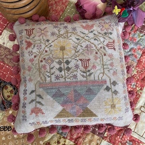 May include: A small, square, cream-colored pillow with a floral embroidery design. The pillow is trimmed with a pink pom-pom border and is sitting on a patchwork quilt with a variety of floral patterns.