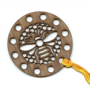 May include: Wooden thread holder with a bee and honeycomb design. The holder has 12 holes for threading and a yellow tassel.