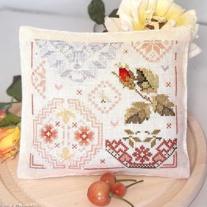 May include: A white pillow with a cross-stitch design featuring a rose, leaves, and geometric patterns in shades of pink, orange, yellow, and blue. The pillow is on a wooden surface with a few red cherries.