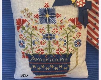 Puede incluir: Una almohada de punto de cruz con un diseño floral azul y blanco en una maceta azul con el texto "Americano!" La almohada está decorada con una cinta azul y un botón rojo y blanco.