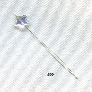 May include: A clear star-shaped bead threaded on a silver beading needle. The needle is straight and has a sharp point. The bead is iridescent and reflects light.