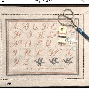 May include: A white linen scissor mat with a cross-stitch design of the alphabet and birds. The mat is surrounded by a black border and includes a pair of scissors, buttons, and thread.