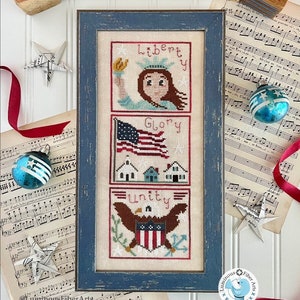 May include: A framed cross-stitch sampler with three panels depicting the Statue of Liberty, the American flag, and an eagle with the words "Liberty", "Glory", and "Unity" respectively. The sampler is surrounded by red, white, and blue decorations, including a star, ribbon, and ornaments.