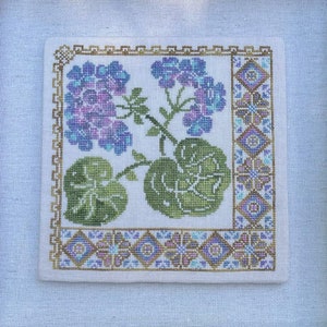 Könnte beinhalten: Eine gerahmte Kreuzstich-Stickerei mit einem floralen Design mit lila Blüten und grünen Blättern. Die Stickerei ist von einem dekorativen Rand mit goldenen, blauen und weißen Akzenten umgeben.