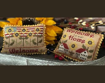 May include: Two embroidered pillow covers with a welcome theme. One features a row of sunflowers with a red, white, and blue background and the text "Welcome". The other features two houses with sunflowers and the text "Welcome to our Home".