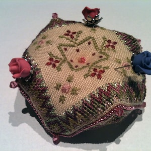 May include: A beige pincushion with a floral pattern and a green border. It has three decorative roses, one pink, one blue, and one red, attached to the top.