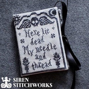 Pode incluir: Um projeto de bordado de ponto de agulha branco com fio preto. O projeto é um pequeno travesseiro retangular com uma fita preta anexada. O travesseiro apresenta um design em preto e branco de uma lápide com o texto "Here lie dead My needle and thread".
