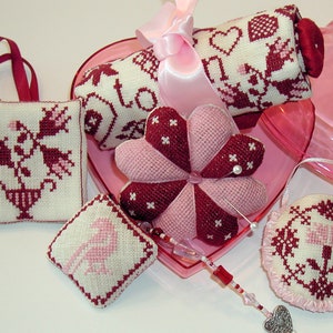 May include: A set of five small, handmade, cross-stitched pillows in pink and burgundy with floral and heart designs. The pillows are displayed in a pink heart-shaped box.