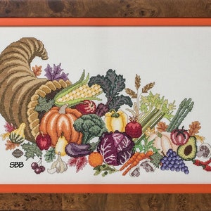 Puede incluir: Obra de arte de punto de cruz de una cornucopia rebosante de verduras y frutas de la cosecha de otoño. El diseño incluye una calabaza, maíz, uvas y otros productos. La obra de arte está enmarcada en un marco de madera con un borde interior naranja.