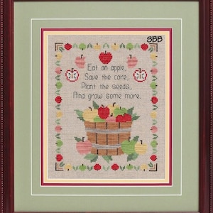 Puede incluir: Bordado de punto de cruz con una cesta de manzanas y el texto "Eat an apple, Save the core, Plant the seeds, And grow some more." El diseño está enmarcado en un marco rojo y verde.