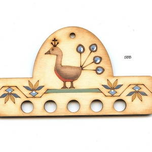 May include: Wooden peacock needle minder with five holes for needles. The peacock is painted in shades of brown, red, and blue. The background is a light wood color with a geometric pattern.