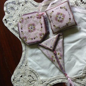 Puede incluir: Una colección de saquitos de lavanda hechos a mano en varias formas: hexagonal, cuadrado y triangular. Cada saquito es de color lila claro, bordado con diseños florales en tonos morados y verdes, y adornado con cuentas. Se encuentran sobre un mantel de encaje blanco.