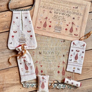 Op de afbeelding: Een verzameling handgemaakte items, waaronder een trekkoordtas, een draagtas en een kruissteeksampler. De items hebben geborduurde ontwerpen in rood en bruin op een crèmekleurige achtergrond. De sampler toont een alfabet en decoratieve motieven.