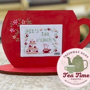 May include: A red wooden teacup with a white cross-stitch design that says "It's tea o'clock". The teacup is on a wooden stand and there is a pink circle with a green teapot and cup that says "Tea Time" and "@xstitchtherainbow".