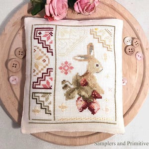 May include: A cross-stitched pillow featuring a bunny holding flowers, with a border of geometric patterns in red, beige, and green. The pillow is on a wooden tray with pink roses and buttons. The text "Samplers and Primitives" is at the bottom.