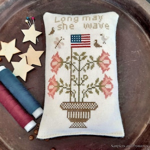 May include: A white cross-stitch sampler with the text "Long may she wave" and a depiction of a potted plant with red flowers and a small American flag.