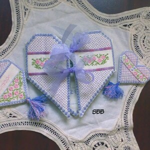 May include: Three handmade heart-shaped fabric ornaments with embroidered floral designs and blue tassels. The ornaments are on a white lace doily. The text "Forever" is embroidered on one of the ornaments.
