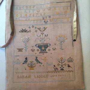 Könnte beinhalten: Ein beigefarbener Stich mit einem Kreuzstich-Motiv, das eine Vase mit Blumen, Vögeln und Pflanzen zeigt. Der Stich ist signiert mit "SARAH LIDDLE aged 12 yrs".