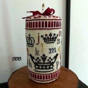 Może przedstawiać: Biały poduszeczka na szpilki z haftem krzyżykowym, z czerwoną i białą kratą na górze i czerwoną wstążką. Poduszeczka jest ozdobiona czarnymi koronami i literami. Poduszeczka leży na drewnianej powierzchni.