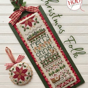 May include: A Christmas Folk cross-stitch design featuring a long, rectangular bookmark and a round ornament. The bookmark has "merry christmas" stitched on it, with festive imagery. The ornament has a poinsettia design. Both have red, green, and cream colors.
