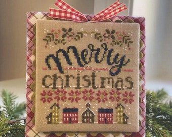 Pode incluir: Um padrão de ponto cruz que representa uma cena de inverno com casas e o texto "Merry Christmas". O padrão é emoldurado e exibido em um pequeno cavalete.