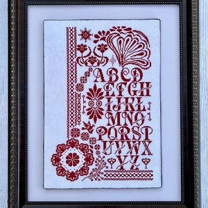 Puede incluir: Una bordadura de punto de cruz enmarcada que presenta un alfabeto rojo con diseños decorativos florales y geométricos. El alfabeto está bordado en un estilo tradicional audaz.