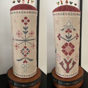 Może przedstawiać: Cylindryczny poduszeczka do szpilek z beżową lnianą poszewką, ozdobioną haftem kwiatowym w odcieniach różowego, czerwonego i szarego. Poduszeczka do szpilek jest zwieńczona burgundowym filcowym poduszkiem i ma drewnianą podstawę z wzorem złotych gwiazd.