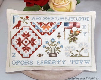 May include: A white pillow with a cross-stitch design featuring the alphabet, a red, white, and blue American flag, a red and white geometric pattern, and a floral motif. The pillow is on a wooden surface.