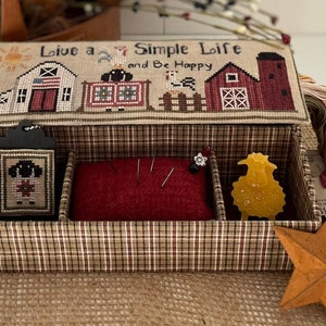 May include: A wooden box with a plaid fabric exterior and a red felt interior. The box has a hinged lid with a cross-stitch design that reads "Live a Simple Life and Be Happy". The box is divided into three compartments, one with a black tag with a cross-stitch design of a sheep, one with a red felt pincushion, and one with a yellow wax sheep.