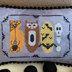 Puede incluir: Una almohada gris con ribete de pompones negros presenta un diseño de punto de cruz de la palabra "BOO" con imágenes de temática de Halloween, que incluyen una araña, un búho, murciélagos y un esqueleto.