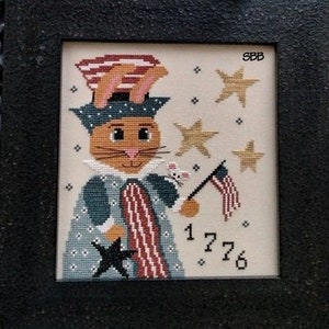 Puede incluir: Un diseño de punto de cruz con un conejito patriótico que lleva un sombrero con una banda roja, blanca y azul. El conejito sostiene una bandera con estrellas y rayas. El diseño incluye estrellas y el número 1776.