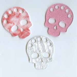 Könnte beinhalten: Drei schädelförmige Garnhalter aus rosa, weißem und rot marmoriertem Kunststoff. Jeder Halter hat sieben Löcher um die Oberseite des Schädels.