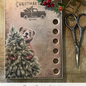 May include: A wooden thread keeper with a vintage Christmas theme. It features a teddy bear holding a Christmas tree, a truck with a Christmas tree in the back, and the text "Fresh Christmas Tree" and "Est. 1853".