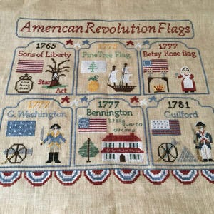 May include: A cross-stitch sampler featuring different flags from the American Revolution. The flags are displayed in a grid format, with the years they were flown. The sampler includes the Sons of Liberty flag, the Pine Tree flag, the Betsy Ross flag, the G. Washington flag, the Bennington flag, and the Guilford flag.