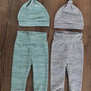 Könnte beinhalten: Zwei Baby-Outfits, jeweils bestehend aus einer geknoteten Mütze und einer passenden Hose. Ein Set ist türkis mit weißen Streifen, das andere grau mit weißen Streifen. Die Outfits werden auf einer Holzoberfläche präsentiert.