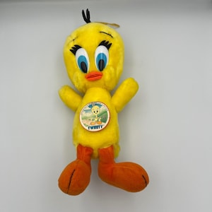 May include: A plush yellow Tweety Bird doll with black eyelashes, blue eyes, and an orange beak and feet. The doll has a round emblem on its chest with a picture of Tweety Bird and the word "Tweety".