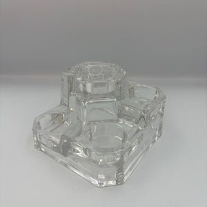 May include: A clear, multi-compartment glass container with a geometric design. The container has several sections, including round and rectangular openings. The glass is transparent, allowing light to pass through. The object is likely a desk organizer or a decorative item.