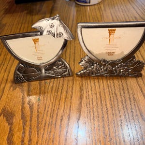 May include: Two silver-toned metal photo frames. One frame has a sun and mountain design, the other has a grape cluster design. Each frame has a semi-circular opening for a 3.5x2.25 inch (9x6cm) photo and the word "CONNOISSEUR".