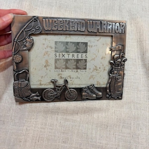 May include: A bronze-colored metal picture frame with the words "WEEKEND WARRIOR" at the top. The frame features embossed images of fishing gear, a bicycle, hiking boots, and golf clubs. The frame has a SixTrees logo.