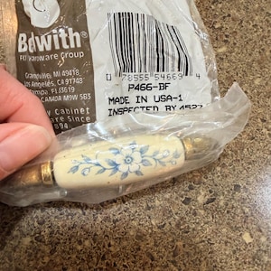 May include: A cabinet pull handle in a clear plastic bag with a label reading Belwith FKI Hardware Group P466-BF. The handle features a cream-colored ceramic center with a blue floral pattern and metallic gold-toned end caps and mounting hardware.
