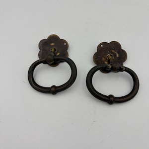 May include: Two antique-style brass drawer pulls featuring a dark, weathered finish. Each pull consists of a circular, flower-shaped backplate with a central hole and a hinged, oval-shaped drop handle that hangs vertically from the mounting point.