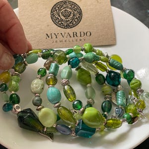 May include: A set of five green glass bead bracelets with silver accents. The bracelets are stacked on a white plate. The image also includes a business card with the text "MyVardo Jewellery".